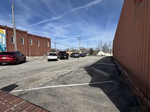 View of an outdoor parking lot with several parked cars, a brick building, and a large orange shipping container under a blue sky with scattered clouds.