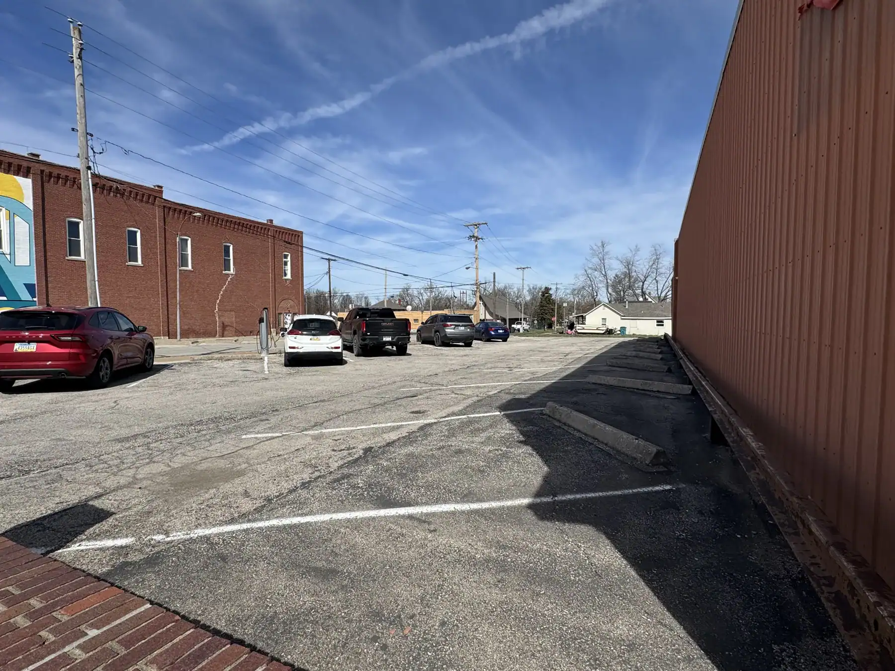 View of an outdoor parking lot with several parked cars, a brick building, and a large orange shipping container under a blue sky with scattered clouds.